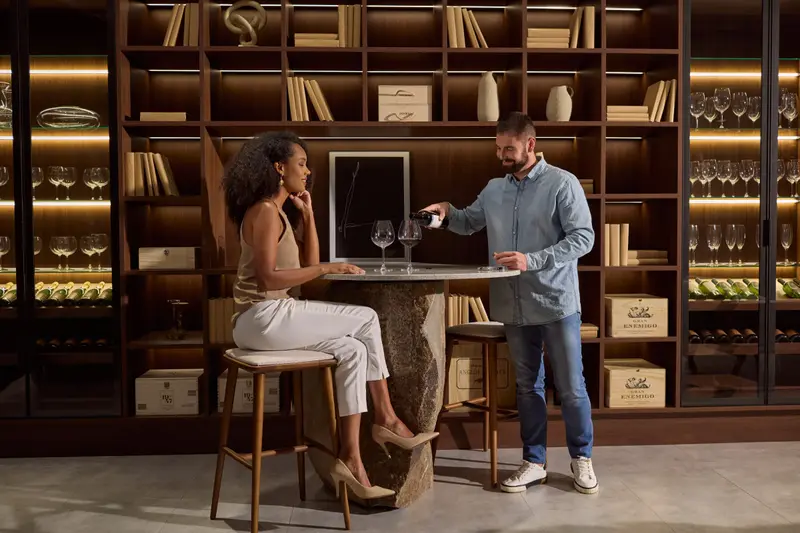 Casal brindando em frente a uma adega e biblioteca planejadas e iluminadas